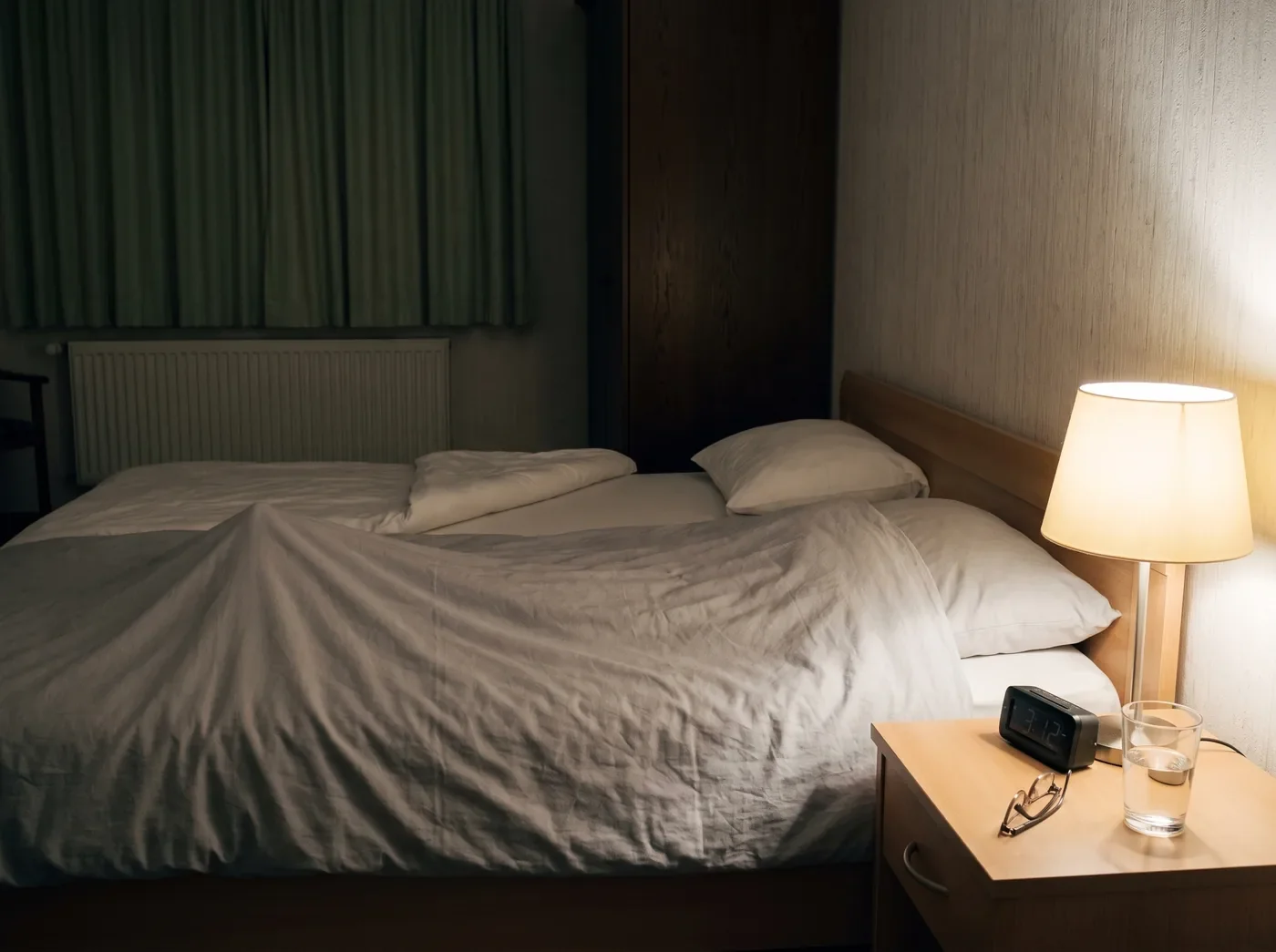 Nachttisch in einem deutschen Schlafzimmer, Wecker zeigt 3:12 Uhr, Nachttischlampe an, im Hintergrund eine Bettdecke, di