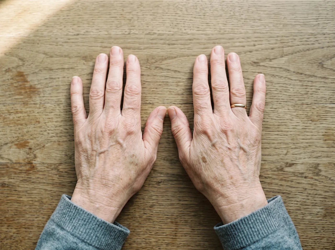 Zwei Hände einer Frau um 60, Handflächen nebeneinander auf einem Holztisch, schlichter Ehering, Tageslicht, als Bild für