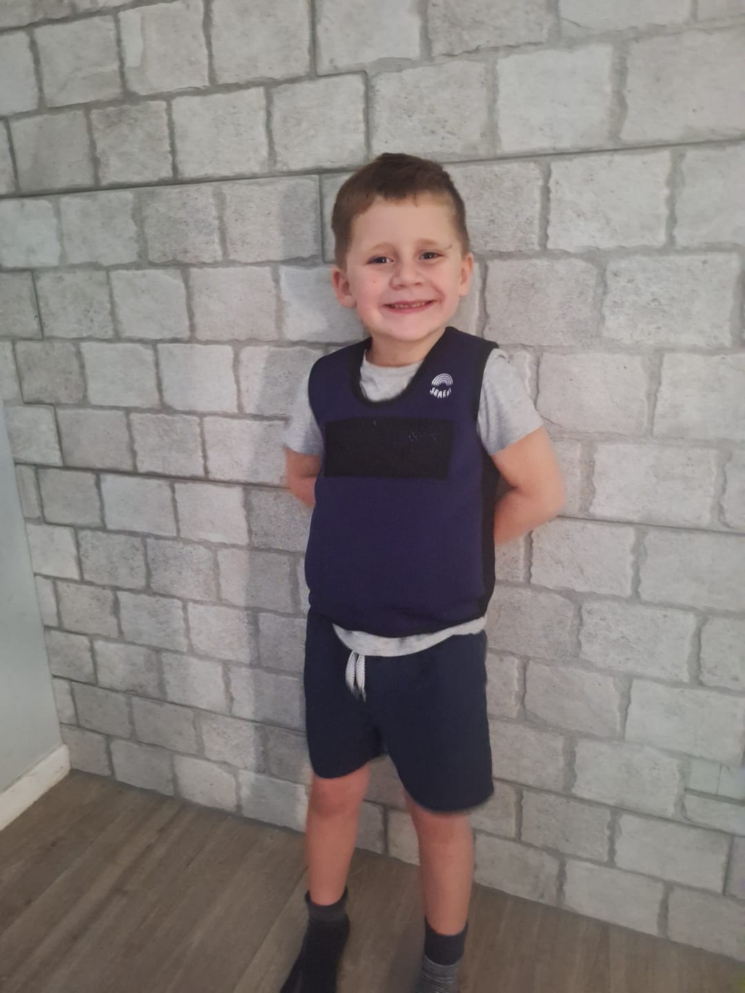 Smiling child wearing a vest, standing against a brick-patterned wall.