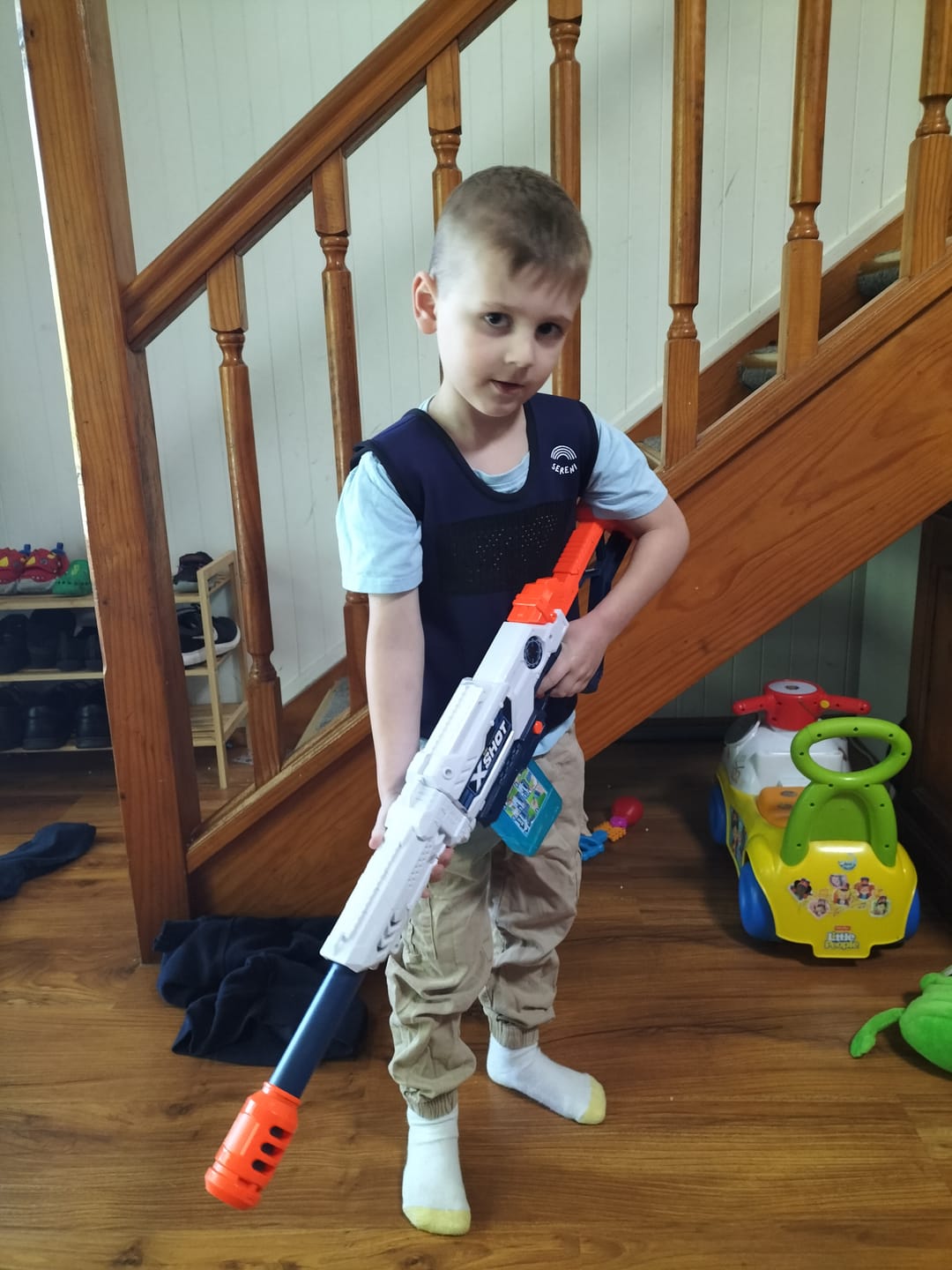 Child with toy blaster stands indoors near staircase.