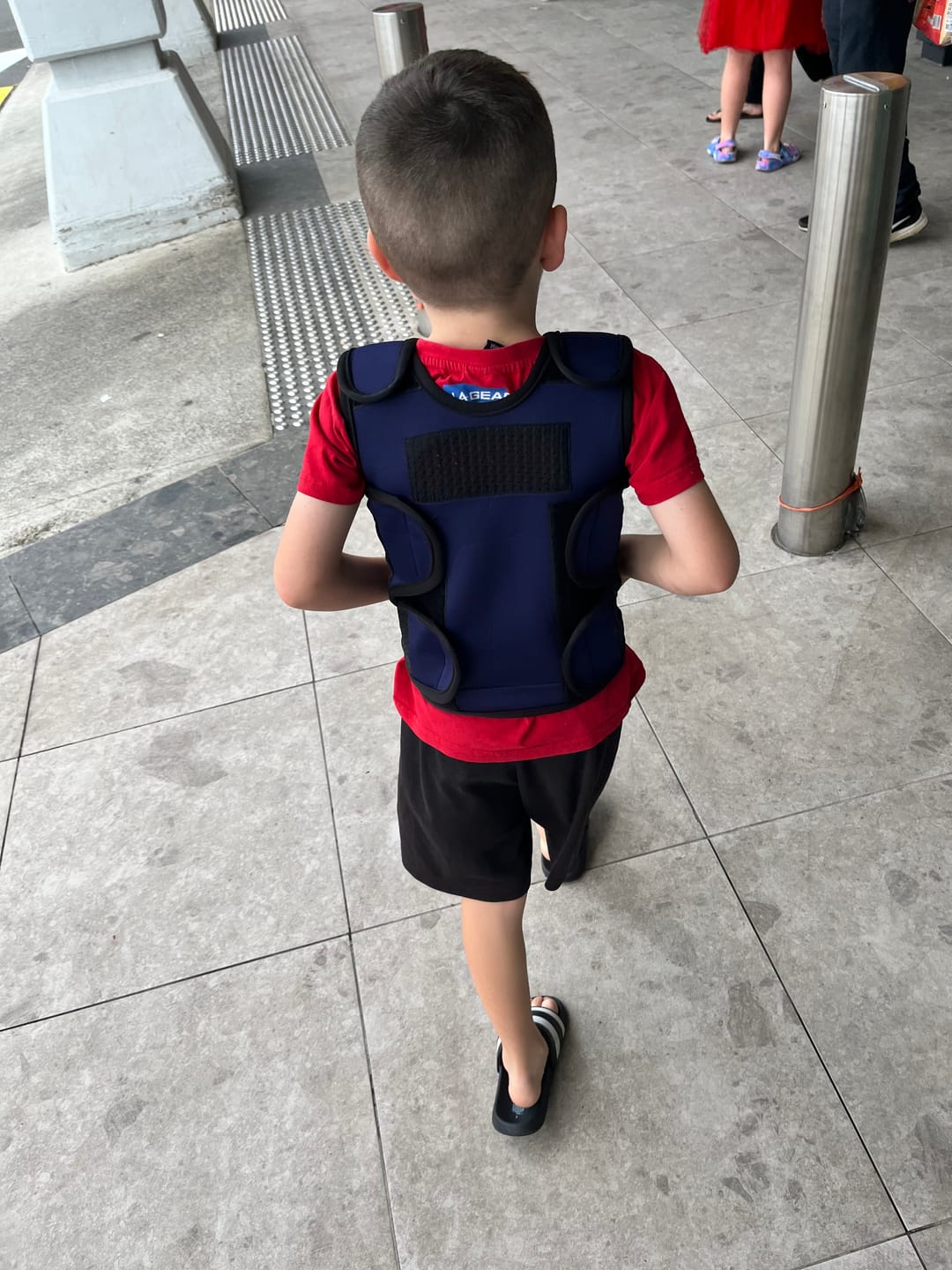 Child wearing a blue vest walks on a tiled sidewalk.