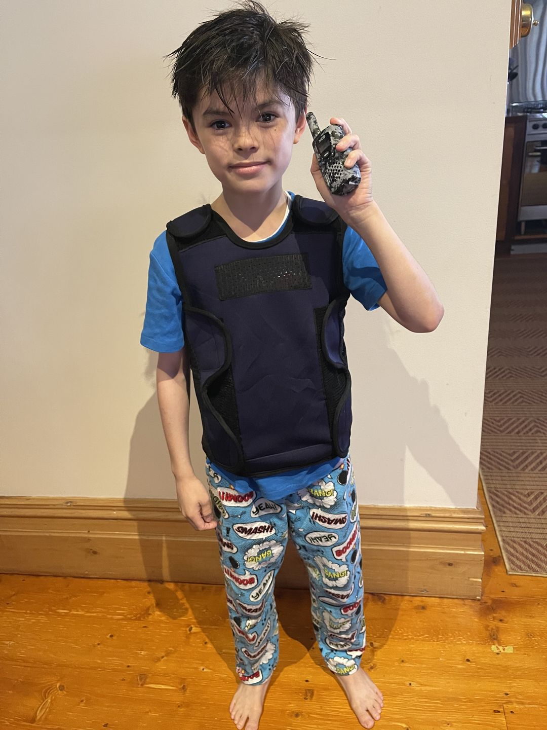 Child in protective vest holding toy walkie-talkie, wearing colorful pajama pants.