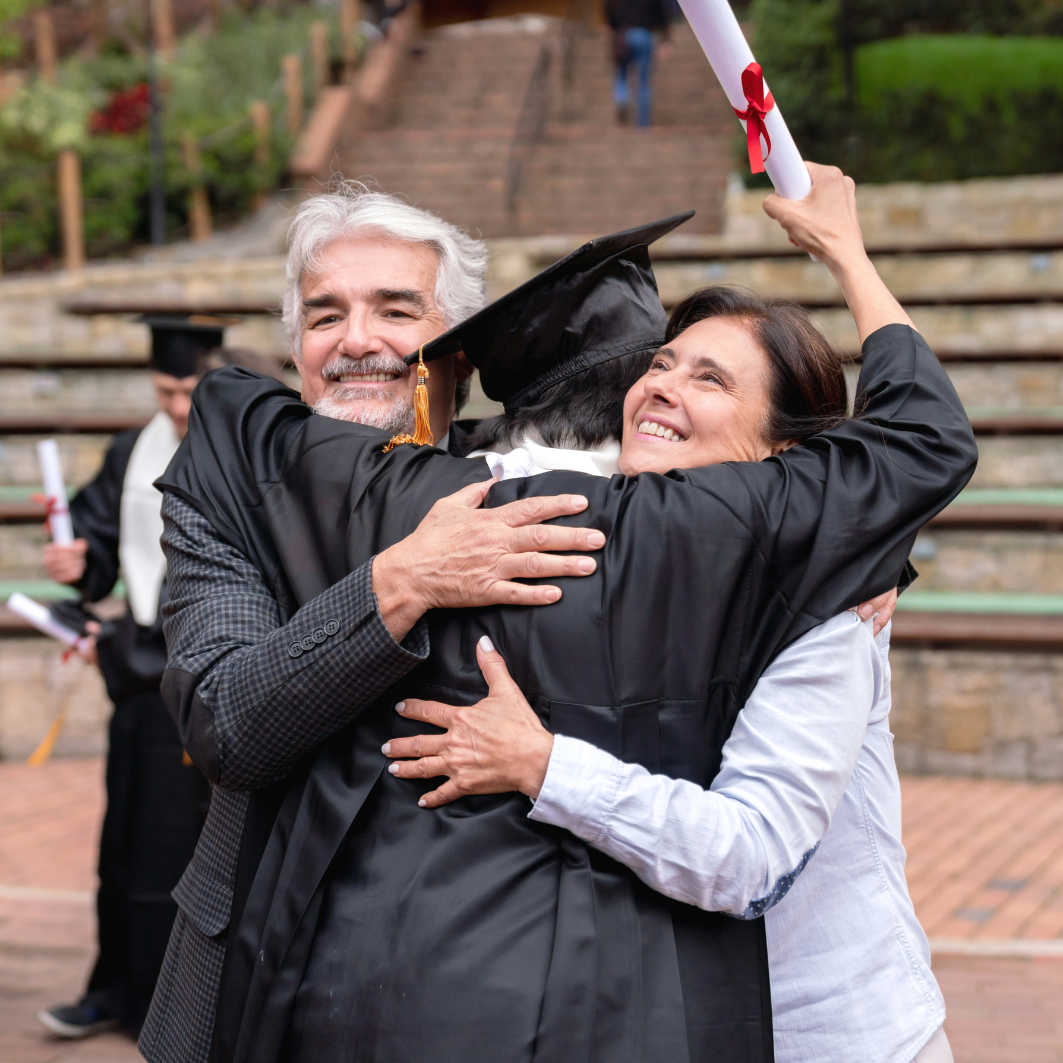 Graduate in cap and gown hugs two people, celebrating graduation outdoors.