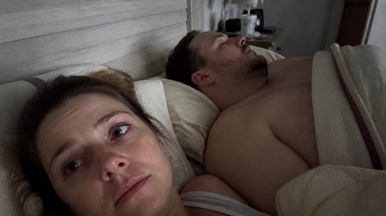 A woman lying awake on her side in bed at night, eyes open, her partner asleep beside her.